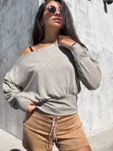 Buzo de tela angorina elastizada color gris, con diseño de cuello bote que permite usarlo con un hombro descubierto. Presenta un corte amplio en la parte superior y una cintura ajustada, con mangas largas.