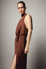 Vestido midi de color marrón con cuello halter y lazada en la nuca. Presenta un diseño con espalda descubierta, frunces en la cintura y una abertura lateral en la falda.