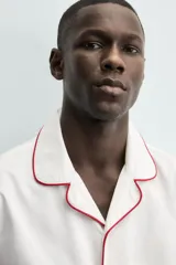 Pijama de dos piezas confeccionado en popelín de algodón blanco, con vivos de ribete en color bordó. Incluye una camisa de manga larga con cuello bowling, bolsillos de plastrón y cierre frontal de botones, y un pantalón de corte relajado con cintura elástica y bolsillos laterales.