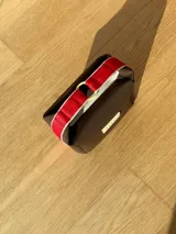 Neceser de neopreno color borgoña, con cierre superior y forro interior. Tiene un asa de mano con una franja azul y blanca.