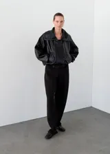 Parka de cuero ovino negro con diseño oversize, cuello embudo con cierre de cremallera, solapas amplias, cierre oculto de cremallera y botones, mangas largas con puños elásticos y bolsillos laterales.