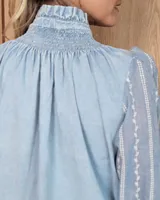 Camisa de denim celeste con detalles de broderie en las mangas, cuello alto con volados y canesú con frunces. Presenta cierre frontal con botones y mangas largas con puños elásticos.