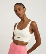 Top deportivo color blanco hueso, de corte ajustado, con breteles anchos y banda elástica inferior con inscripción. Presenta un pequeño recorte circular en el centro del busto.