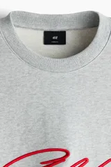 Buzo de cuello redondo en tejido de algodón gris melange, con diseño de hombros caídos y bordado frontal con la palabra 'Golden' en color rojo. Presenta puños y bajo acanalados.