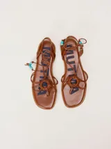 Sandalias planas de cuero color marrón, con diseño de punta cuadrada, tiras finas cruzadas y un detalle trenzado circular en el empeine. Cuentan con tiras para atar al tobillo decoradas con cuentas turquesas en las puntas.