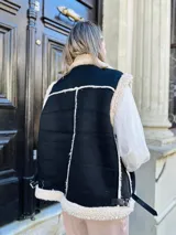 Chaleco de gamulán negro con interior y bordes de corderito sintético color crema. Presenta cierre frontal y bolsillos laterales con cierre.