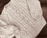 Sweater artesanal de tejido de punto de lana merino color marfil, de calce amplio y relajado, con cuello redondo y diseño frontal en relieve con trenzas y bodoques. Presenta mangas largas abullonadas con paneles texturizados y puños acanalados.