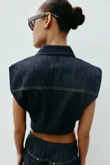 Chaleco de denim oscuro con cuello solapa, cierre frontal con cremallera metálica y bolsillos delanteros con tapeta. Presenta un diseño de corte abullonado con bajo elástico.