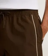 Bermuda deportiva color marrón con piping en color crema, confeccionada en poliamida de secado rápido. Cuenta con cintura elástica con cordón ajustable y bolsillos laterales.