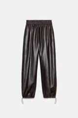 Pantalón de tiro alto confeccionado en material con efecto piel, con cintura elástica, bolsillos delanteros y nervio marcado en el frente. Presenta un diseño bombacho con bajo ajustable mediante cordones elásticos.