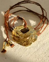 Collar artesanal con dije de flor en bronce o alpaca, suspendido en un cordón de seda natural teñido a mano. El diseño permite atarse con un moño, ofreciendo versatilidad para lucirlo de distintas formas.