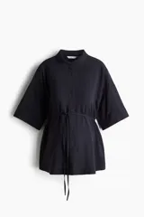 Camisa negra de viscosa liviana, con cuello clásico, botonadura frontal, hombros caídos y mangas cortas hasta el codo. Cuenta con un cordón ajustable en la cintura y bajo recto.