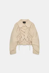 Chaqueta corta color beige, confeccionada en gabardina, con cuello solapa redondeado, mangas largas con frunces y detalle de frunces con lazada en el delantero, cerrando con botones.