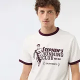Camiseta blanca de manga corta con ribetes en contraste de color bordó en el cuello y los puños. Presenta un estampado gráfico frontal con la inscripción "Stephen's Running Club Est. 88 Sacramento" y la silueta de un corredor.