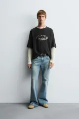 Camiseta de punto de algodón, corte boxy fit, color negro con efecto lavado. Presenta un estampado gráfico frontal que simula un ojo o forma ovalada con texto ilegible en su interior.