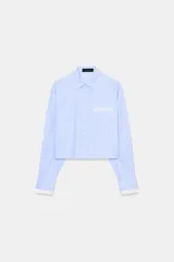Camisa de manga larga, corta (crop), con cuello solapa y bolsillo de vivo en el delantero. Presenta un patrón de rayas verticales azules y blancas, con detalles de acabado en blanco en los puños y el bolsillo.