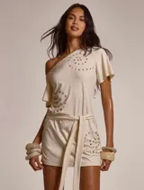 Vestido corto de color beige con diseño asimétrico de un solo hombro y manga corta, adornado con patrones circulares de botones decorativos. Incluye un lazo ajustable para la cintura.