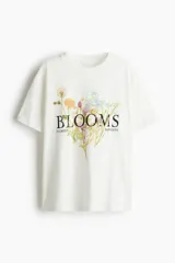 Remera de corte oversized confeccionada en punto de algodón, con cuello redondo ribeteado, mangas cortas y bajo recto. Presenta un estampado frontal con motivos florales y la palabra 'BLOOMS'.