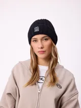 Gorro tipo beanie de punto acanalado en color lila, con parche rectangular negro en el frente que incluye la inscripción 'EAT THE WAVE'.