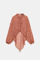 Camisa de estampado floral con cuello subido, lazo frontal y mangas largas con puños elásticos. Presenta un diseño con bajo asimétrico y cierre posterior con botones ocultos.