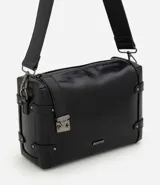 Cartera tipo shoulder de tamaño mediano, confeccionada en poliuretano con diseño rectangular. Presenta correas laterales decorativas con remaches metálicos, cierre superior con cremallera, herrajes en acabado gunmetal y correa larga ancha ajustable.