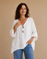 Blusa de lino color blanco, de corte amplio y holgado, con escote en V decorado con dos botones grandes y cordones rústicos colgantes. Presenta mangas 3/4 con presilla y bolsillos delanteros aplicados con detalles de flecos.