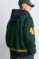 Cazadora tipo varsity de corte relajado confeccionada en tejido efecto ante color verde oscuro. Presenta cuello solapa, cierre frontal con botones a presión y puños y bajo acanalados con rayas en contraste. Incluye parches bordados en color amarillo en el frente y mangas.