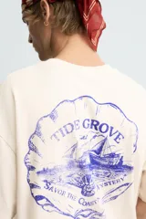 Camiseta de corte boxy fit, color beige con efecto lavado, cuello redondo y manga corta. Presenta un estampado gráfico en azul oscuro en el frente con tipografía estilo vintage.