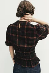 Camisa de cuello redondo y manga corta abullonada, con diseño de cuadros en tonos negro y marrón. Presenta un cuerpo entallado mediante frunces elásticos en la cintura y cierre posterior con abertura y botón.