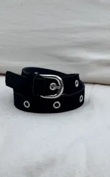 Cinturón de gamuza color marrón con ojales metálicos plateados distribuidos a lo largo de la tira y hebilla metálica de diseño redondeado.