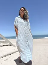 Vestido camisero midi de corte holgado, con estampado de rayas verticales celestes sobre fondo blanco. Presenta cuello camisero, cierre frontal con botones, mangas cortas con detalle de trabilla y bolsillos laterales.
