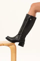 Bota estilo biker de cuero negro con caña alta y cierre metálico lateral. Presenta un diseño versátil con estribo y cadena desmontables, taco cuadrado y suela con plataforma.