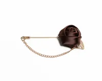 Pin para ojal con diseño de rosa de satén en color marrón, montado sobre una aguja metálica dorada con cadena decorativa y detalle de hoja metálica.