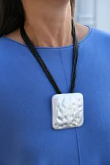 Collar de diseño moderno con dije cuadrado de superficie irregular y acabado plateado mate, suspendido de múltiples cordones negros finos.