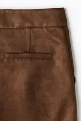 Pantalón de corte bootcut confeccionado en gamuza color marrón. Presenta cierre frontal con botón oculto, bolsillos laterales al bies y costuras decorativas horizontales sobre las rodillas.