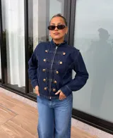 Chaqueta de denim azul oscuro con estilo militar, cuello alto, cierre frontal con cremallera y detalles de trabas decorativas con botones dorados.
