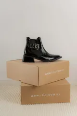 Botineta de cuero charol negro con acabado brillante, diseño de punta fina y taco cuadrado. Presenta un panel elástico lateral y una correa decorativa con hebilla metálica plateada.
