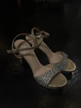 Sandalias de fiesta con plataforma, color dorado, con taco cuadrado alto y tira frontal decorada con pedrería brillante. Cuenta con pulsera ajustable al tobillo con hebilla metálica.