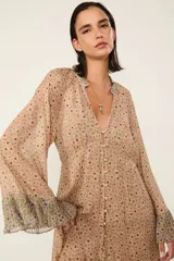 Vestido largo beige estampado con diseño floral, con forro, escote en V con cierre de botones delanteros y mangas largas con puños anchos con volados. Presenta una silueta fluida, detalles plisados y un volante superpuesto en el bajo.