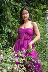 Vestido largo de fiesta color fucsia, sin mangas y con escote strapless. Presenta un corte a la cintura y falda con vuelo. El corpiño tiene un diseño drapeado con abertura en el centro.