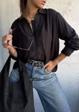 Camisa negra de corte holgado con detalles de encaje floral en la parte frontal y en los puños, cierre frontal con botones y cuello clásico.