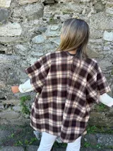 Ruana estilo chaqueta con diseño de cuadros escoceses en tonos marrones y beige. Presenta un cierre frontal tipo alamar con detalle en cuero sintético y bordes terminados.