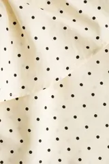Blusa de viscosa color beige con estampado de lunares negros. Presenta cuello con volados, cordón fino para ajustar, botonadura frontal y mangas largas con puños elásticos fruncidos.