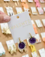Aritos tipo argolla de acero con dije circular color violeta, enmarcado por una hilera de cristales brillantes.