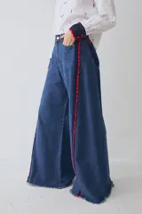 Jean de corte wide leg en color azul denim, con detalle de apliques de crochet en color rojo y azul a lo largo de las costuras laterales frontales. Presenta ruedo deshilachado y cierre frontal con botón metálico.