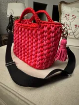 Cartera tejida a crochet con hilo de algodón en tonos rosa y rojo. Tiene doble asa corta y correa larga de tela negra regulable. Incluye un dije de borla rosa.