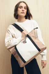 Bolso tipo shopper de nylon, con diseño bicolor en tonos beige y blanco. Cuenta con asas largas de cinta negra, un bolsillo frontal contrastante y detalle de tiras decorativas con cuentas en el cierre.