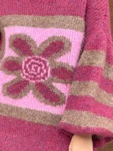 Buzo de tejido de punto con textura peluda, de corte holgado y mangas amplias. Presenta un diseño geométrico con una flor grande estampada en el centro, combinando tonos fucsia, rosa y marrón en un patrón de bloques y rayas.