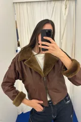 Campera bomber corta de cuero ovino marrón con detalles en napalan coñac y forro interior. Presenta cuello solapa, cierre frontal con cremallera de doble sentido, bolsillos laterales ribeteados, mangas largas con detalles de pasamanería cruzada y puños de piel sintética.