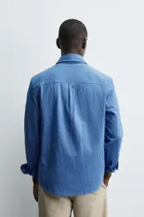 Camisa de corte regular fit confeccionada en tejido de algodón con textura. Presenta cuello solapa, cierre frontal de botones, manga larga con puños abotonados y un bolsillo plastrón en el pecho.
